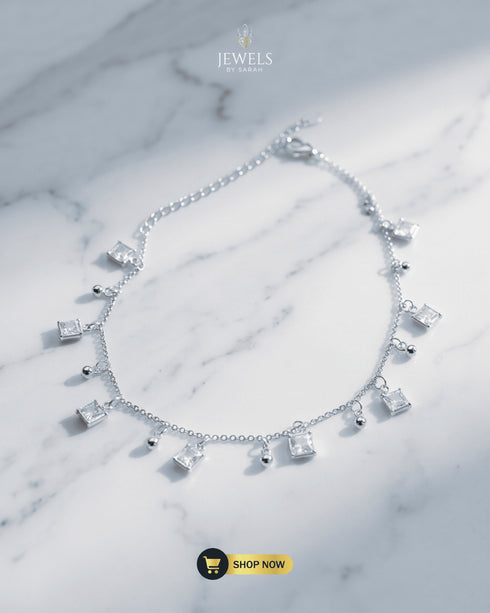 Elegant silver anklet with princess-cut square Zircon charms and silver beads displayed on a marble surface.