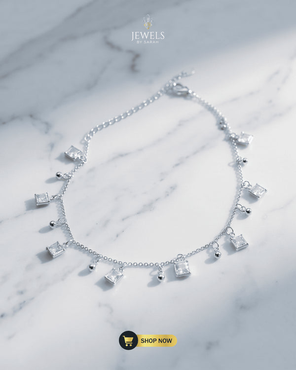 Elegant silver anklet with princess-cut square Zircon charms and silver beads displayed on a marble surface.
