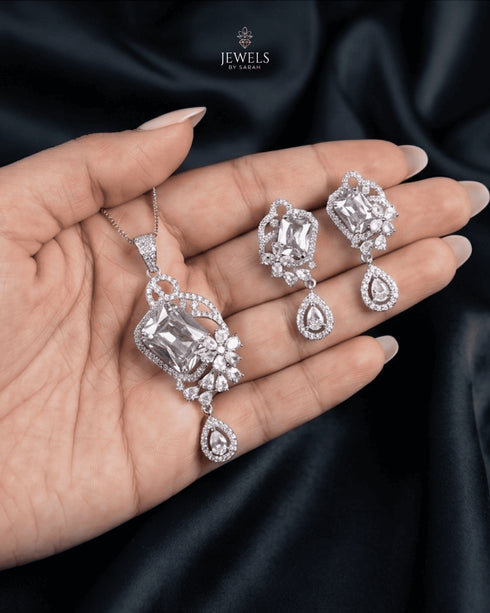 Aura luxury emerald-cut crystal clear pendant set with matching drop earrings held against a dramatic black silk background.