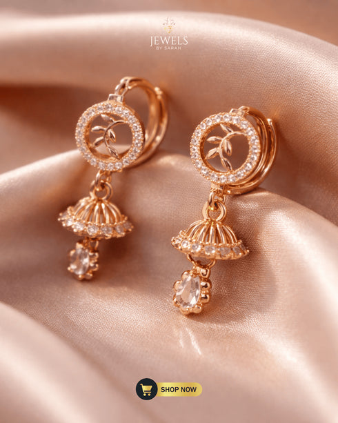 Shimmering rose gold zircon jhumka earrings shown at an angle on a soft champagne silk background.