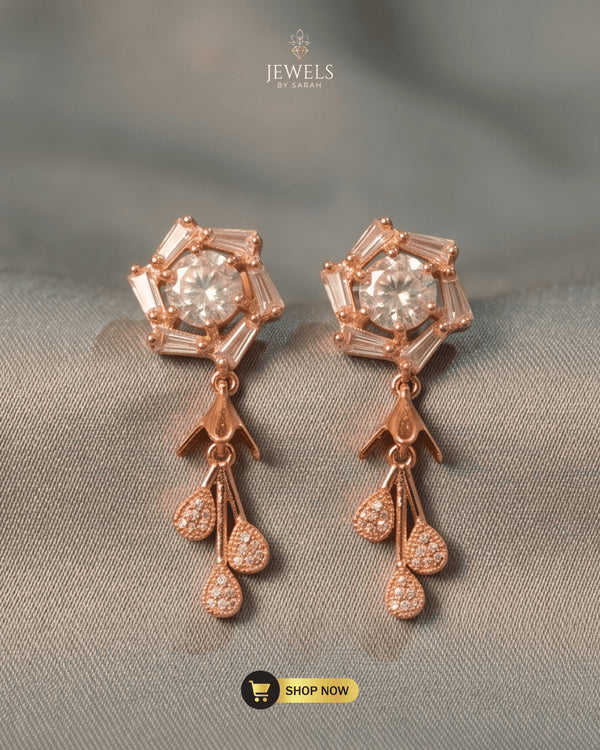Pair of rose gold geometric blossom earrings with Zircon baguette crystals and teardrop tassels on a textured grey background.