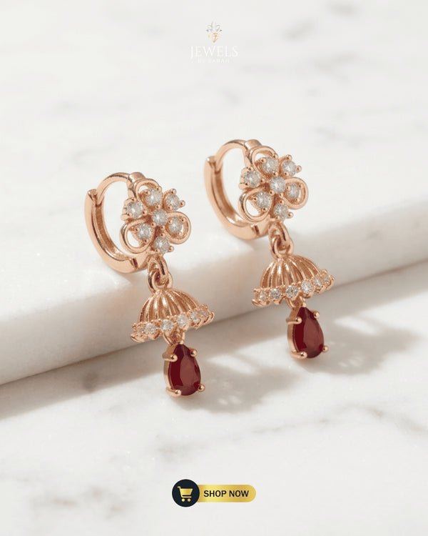 Pair of rose gold floral jhumka huggie earrings with ruby red Zircon drops on a white marble surface.