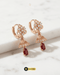 Pair of rose gold floral jhumka huggie earrings with ruby red Zircon drops on a white marble surface.