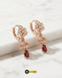 Pair of rose gold floral jhumka huggie earrings with ruby red Zircon drops on a white marble surface.