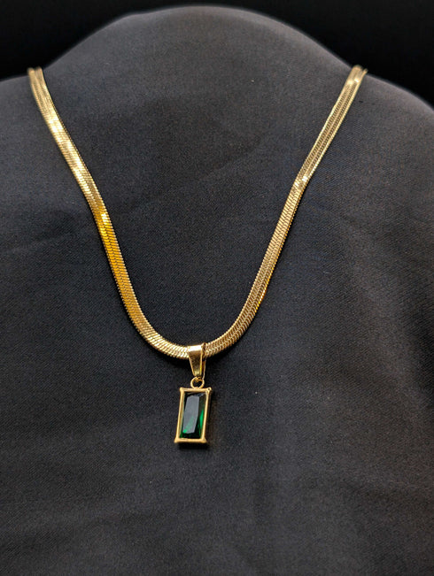Gold-plated stainless steel herringbone chain necklace with a rectangular emerald green baguette stone pendant against a black background.