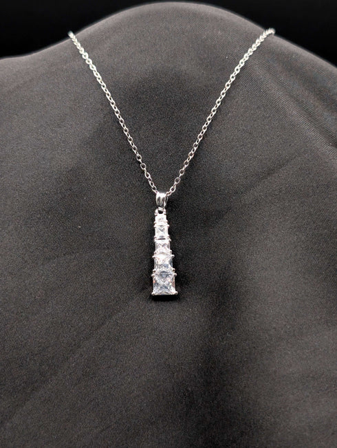 Silver-toned stainless steel necklace with a vertical tiered drop pendant featuring five graduated square-cut crystals on a black background.