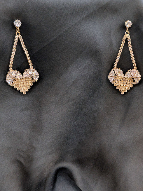 Gold-toned chandelier earrings with pave crystal heart pendants and triangular drop accents against a dark background.