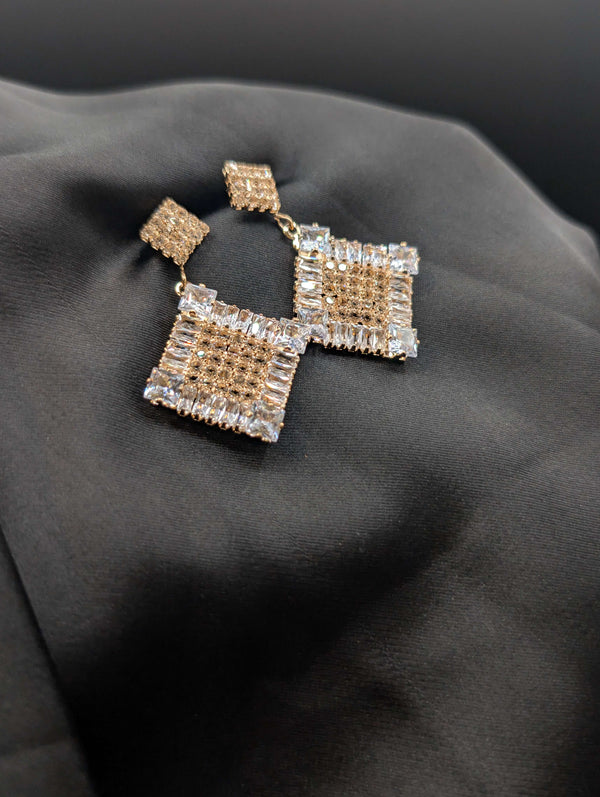 Gold-toned geometric statement earrings with square drop pendants featuring champagne and clear crystal pave on a dark background.