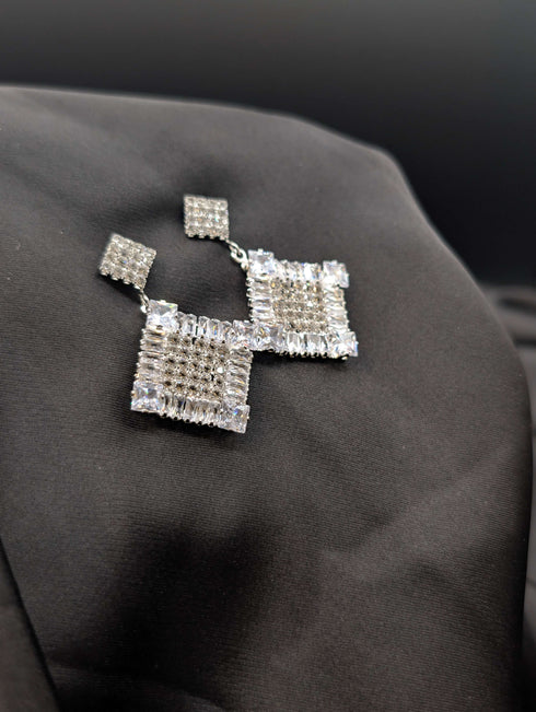 Silver-toned geometric statement earrings with large square drop pendants featuring clear crystal pave on a black background.