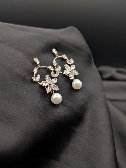 Gold-toned drop earrings featuring a marquise crystal flower blossom design and a white pearl drop on a dark fabric background.