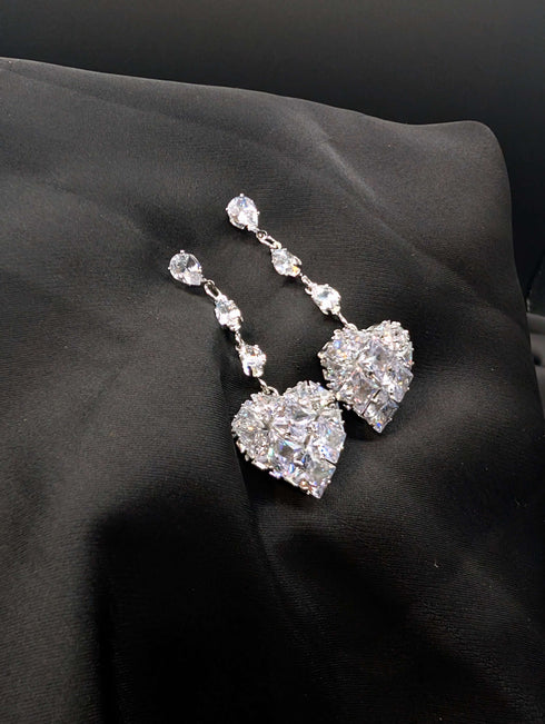 Silver-toned statement drop earrings featuring three pear-cut crystals and a large pave-encrusted heart pendant on a dark background.
