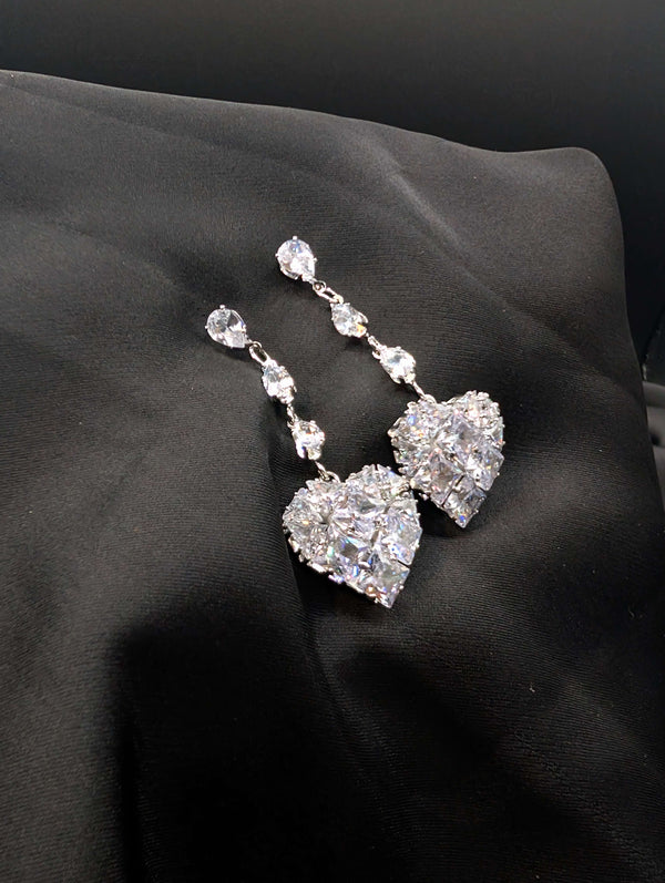 Silver-toned statement drop earrings featuring three pear-cut crystals and a large pave-encrusted heart pendant on a dark background.