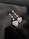 Silver-toned statement drop earrings featuring three pear-cut crystals and a large pave-encrusted heart pendant on a dark background.