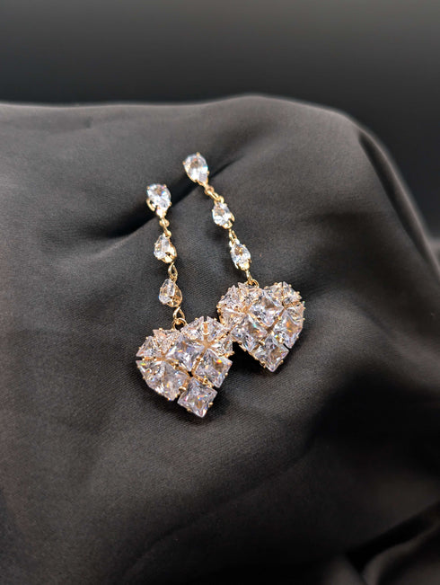 Gold-toned statement drop earrings featuring a vertical row of pear-cut crystals and a large pave heart pendant on a dark background.