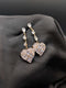 Gold-toned statement drop earrings featuring a vertical row of pear-cut crystals and a large pave heart pendant on a dark background.