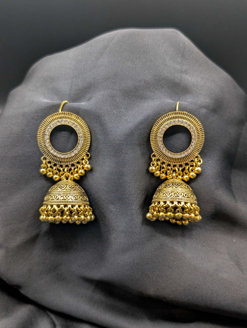 Antique gold-toned Jhumka earrings featuring a crystal-encrusted circular stud and detailed filigree bell pendants with gold bead accents on a dark background.