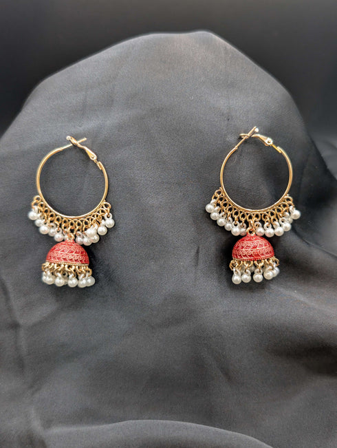 Gold-toned Jhumka hoop earrings with vibrant ruby red enamel bells and white pearl fringe against a dark background.