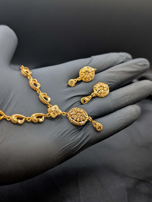 Handheld view of a 24k gold plated necklace with heart-shaped links and matching floral drop earrings against a dark background.