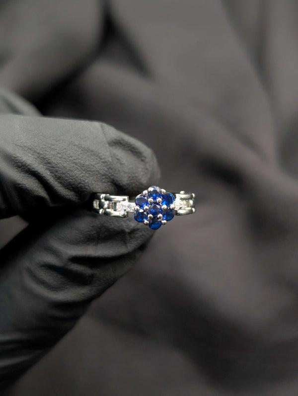 Silver ring featuring a blue sapphire-colored flower cluster center and a unique articulated link band held against a black background.