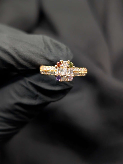 Gold statement ring featuring a rectangular clear baguette-cut center surrounded by multi-colored round gemstones.