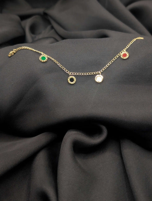 Gold link chain bracelet featuring five circular dangling charms with green, red, black, and white centers laid across a dark grey textured fabric.