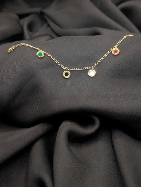 Gold link chain bracelet featuring five circular dangling charms with green, red, black, and white centers laid across a dark grey textured fabric.