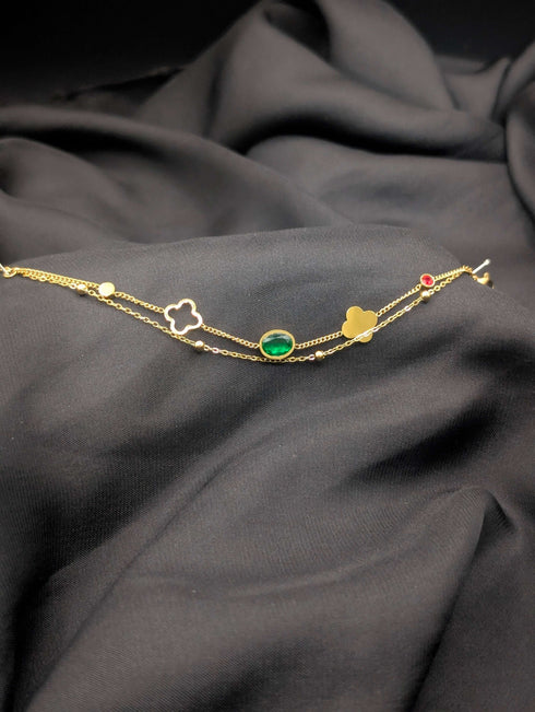 Gold-tone double-strand bracelet with an oval emerald green gemstone, gold clover charm, and small red crystal accent laid on black fabric.