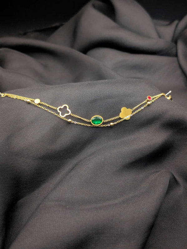 Gold-tone double-strand bracelet with an oval emerald green gemstone, gold clover charm, and small red crystal accent laid on black fabric.