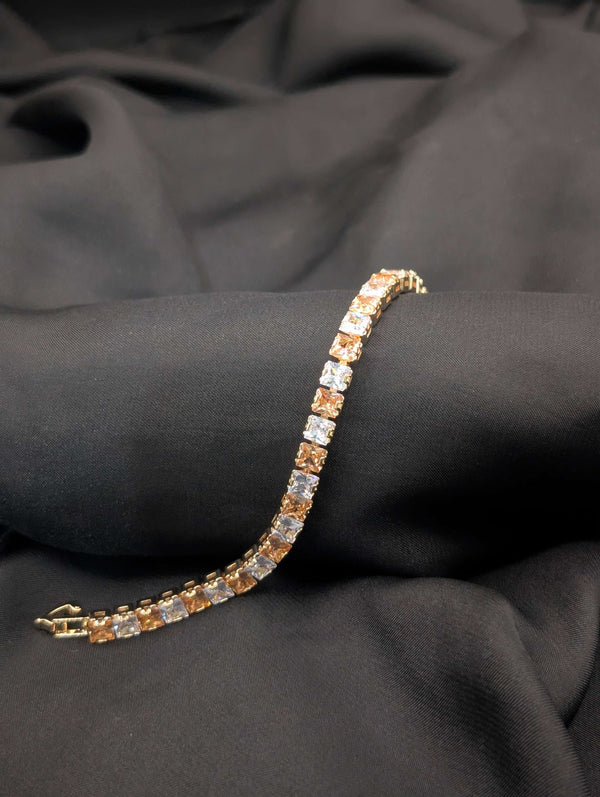 Gold-tone tennis bracelet with alternating champagne and clear princess-cut crystals draped across a black textured background.