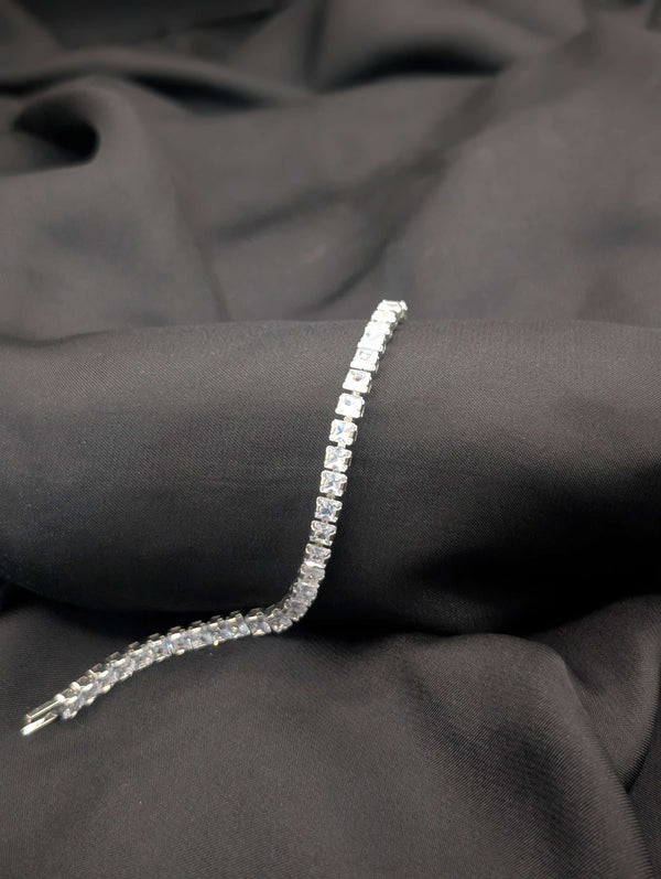 A classic all-clear princess-cut tennis bracelet with a polished silver-tone finish draped across a textured black fabric.