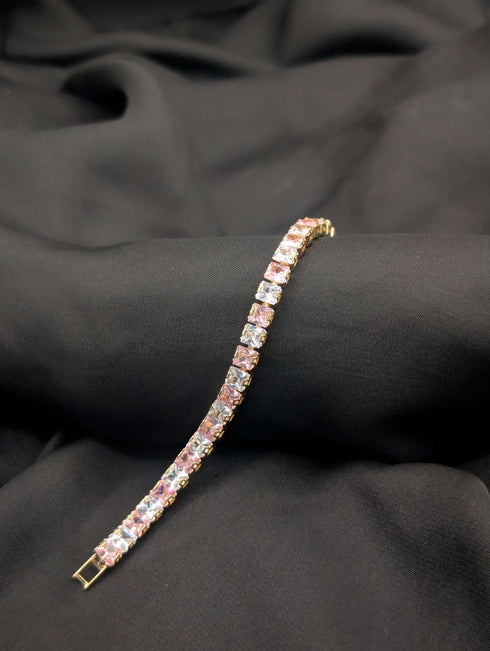 Polished gold-tone tennis bracelet with alternating blush pink and clear white princess-cut crystals on a dark textured background.