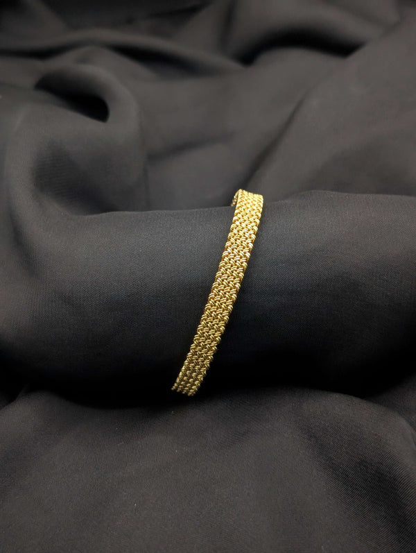 A polished gold-tone bangle with an intricate woven mesh texture laid across a dark, draped fabric.