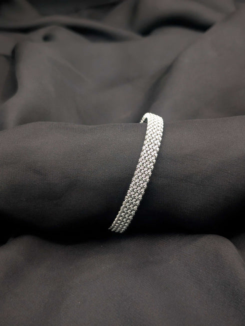 A polished silver-tone bangle with an intricate woven mesh texture displayed on a dark, flowing fabric background.