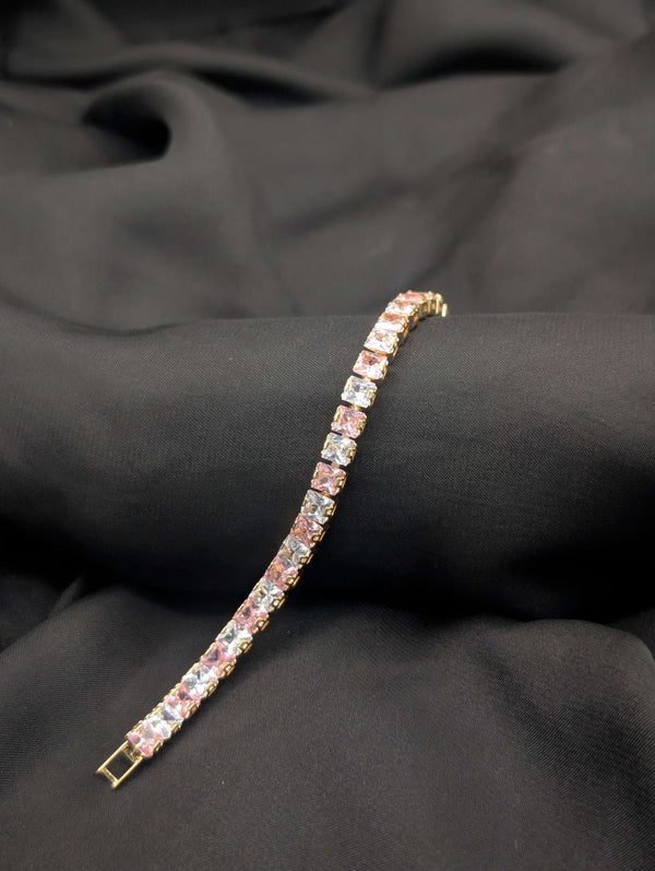 Polished gold-tone tennis bracelet with alternating blush pink and clear white princess-cut crystals on a dark textured background.